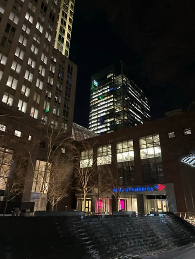 The nighttime image features several buildings, including "Bank of America" at Founders Hall. A modern skyscraper with illuminated windows stands at an angle against the dark sky. Another building with numerous illuminated windows is visible in the lower left. A dark, textured structure, potentially a water feature, is present on the right edge of the frame, and bare tree branches are visible in the foreground.