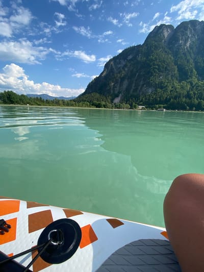The image shows a person on a paddleboard on a calm, turquoise lake. The paddleboard is white with orange diamond patterns and has a black bungee cord attachment. The person's knee and elbow are visible at the top right of the frame. In the background, a steep, tree-covered mountain rises from the shore of the lake. The sky is bright blue with scattered white clouds. The scene appears to be set in a mountainous region during daylight, likely in a natural setting such as a lake in the Alps, possibly in Austria, given the location context provided. The overall mood is peaceful and recreational, suggesting a day of paddleboarding. No visible text is present in the image.
