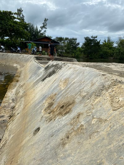 The image depicts a concrete dam structure situated in the city of Aneuk Paya, Indonesia. On the dam's walkway, three young males are visible. Two of them are facing away from the camera, one in a green t-shirt and the other in a white t-shirt. The third individual, wearing a black t-shirt and shorts, is also facing away from the camera but appears to be looking towards the right side of the frame. To the left of the dam, several motorcycles are parked alongside a row of trees. In the background, a rustic wooden structure with a dark roof is nestled among dense green foliage. The sky is overcast with heavy clouds, suggesting a potentially rainy or gloomy day. The dam itself is constructed of rough concrete, showing signs of wear and tear with stains and some discoloration. Water is visible on the left side of the dam, indicating a body of water it is holding back. The overall scene suggests a casual outdoor setting in a rural or semi-rural area.