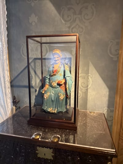 A framed glass display case containing a statue of a seated Asian man wearing a robe is prominently featured. The statue, dressed in teal and gold, is placed atop a dark, ornately carved wooden chest, the surface of which is partially covered with a clear protective layer. The backdrop is a textured, light blue wall decorated with a repeating floral motif. The scene is indoors. The image was captured inside the Pena National Palace in Sintra, Portugal.