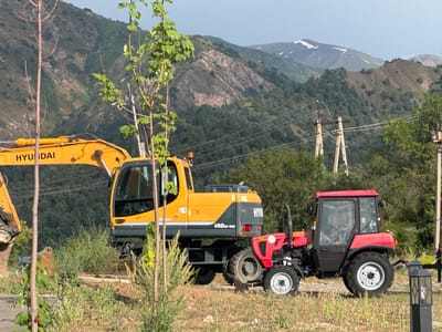 In the city of Uchterek, Uzbekistan, a bright yellow Hyundai excavator is parked next to a red tractor. The excavator, a Hyundai 140w-9S, has its arm extended and is positioned on a slight incline. The tractor, a Belarus 4221, is smaller and sits to the right of the excavator. Both machines are stationary. In the background, lush green trees cover the slopes of rolling mountains, and power lines crisscross the scene. The sky is a pale blue with some light clouds. The overall environment appears to be a rural or semi-rural construction or work site.