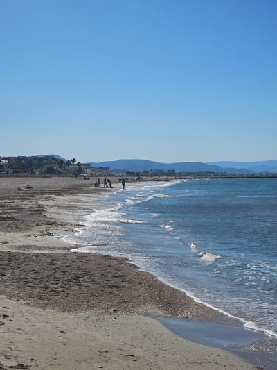 This is a medium shot, eye-level, vertical photograph taken at Deveses, Spain, capturing a seaside scene on a clear day. The image is dominated by a vast, unblemished blue sky that occupies the upper half. Below the sky, a tranquil, azure sea stretches across the frame, with gentle waves lapping at the shore. The water is clear and reflects the bright sunlight, creating sparkling patterns on its surface.
The foreground features a sandy beach, a mix of fine sand and small pebbles. The beach is mostly uncrowded, with a few individuals scattered along its expanse. Closer to the water's edge, a group of people, possibly a family, are gathered. One person is walking along the shoreline, while others appear to be interacting or relaxing. Further back, a lone individual is reclining on the sand.
In the middle ground, a line of buildings, characteristic of a coastal town, runs parallel to the beach. These structures vary in height and style, with some appearing to be hotels or residential apartments, interspersed with palm trees. Beyond the buildings, a range of blue-hued mountains forms a distant backdrop, adding depth to the landscape. The overall impression is one of a peaceful, sunny day at a popular beach destination.
