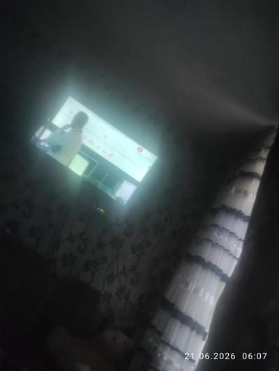 A dimly lit room is captured, with a television screen displaying a person and some graphics. The screen is the primary light source, casting a glow on the surrounding area. The room appears to have patterned wallpaper and a curtain on the right side of the image. A timestamp at the bottom right corner reads "21.06.2026 06:07". The location is Zarafshan City, Uzbekistan.