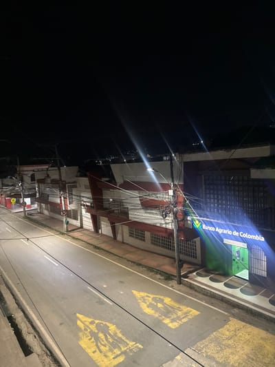This image depicts a nighttime street scene in Timaná, Colombia, viewed from an elevated perspective. The sky is dark and clear, indicating nighttime conditions. A two-lane asphalt road runs horizontally through the midground, illuminated by overhead streetlights that produce visible lens flares. The road surface features two faded yellow pedestrian crossing markings depicting silhouetted figures, likely indicating a school zone, near a white double-center line.

Along the right side of the street, a row of multi-story buildings, primarily white with red accents, features multiple windows protected by security bars. The building on the far right displays a prominent, illuminated sign for "Banco Agrario de Colombia" in blue and green, with a visible green ATM vestibule to its right. A utility pole heavily laden with multiple bundles of black cables stands directly in front of the bank, extending wires across the street.

On the left side of the street, beyond the road, a sidewalk fronts similar buildings. Traffic signage is visible: a red circular "No Parking" sign, a yellow diamond-shaped "School Zone" sign depicting two children, and a white circular speed limit sign (likely 20 km/h). No individuals or vehicles are present in the immediate foreground or midground. Distant hills are faintly visible against the dark horizon in the background.