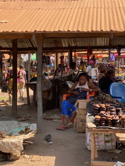 This image depicts an open-air market scene in Nev, Nigeria, under a bright, sunny sky. The ground consists of dry, reddish-brown soil with visible debris.

In the foreground, a woman is seated on an orange plastic chair, facing slightly left. She wears a white tank top, a dark garment across her lap, and blue shorts patterned with white stars and circles. Her bare feet are on the ground, with red flip-flops nearby. She is holding a small, light-colored object in her hands. To her right, a wooden table displays numerous stacks of dried, dark-colored items, consistent with smoked fish or meat products. Behind this table, a blue plastic chair and a cardboard box are visible. To the far left foreground, a white tarp or fabric is spread on the ground, displaying a small pile of green matter.

The market structures are characterized by wooden posts supporting corrugated metal roofs, providing shade. In the midground and background, multiple individuals are present, engaging in various market activities. Several people, including women with headwraps and children, are standing or walking. Some figures are tending to market stalls, where goods like clothing are displayed hanging. The overall environment appears active, with goods spread on tables and ground coverings. The background reveals additional market stalls and distant structures under similar roofing.