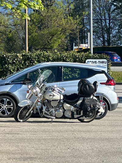 A silver motorcycle with black leather saddlebags and a windscreen is parked next to a light blue Chevrolet Bolt electric vehicle. The motorcycle is positioned closer to the camera than the car. A "Drive-Thru at the..." sign is partially visible next to the car. The scene appears to be in a parking lot next to a hedge. The precise location within Pitt Meadows, Canada, cannot be determined from the visual data.