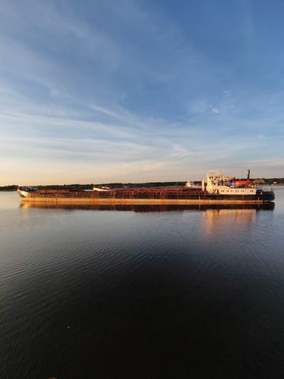 A long cargo ship is sailing on a calm body of water. The ship has a long, flat deck and a superstructure at the rear. The sky above is a clear blue with a few wispy clouds. The sun is setting, casting a warm glow on the ship and the water. The shoreline in the background is lined with trees. The image was taken in Shchetinino, Russia.