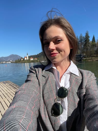 A selfie photograph shows a woman on a wooden dock, likely on Lake Bled in Slovenia, evident from the island church visible in the background. She is wearing a white button-down shirt and a gray plaid blazer. Sunglasses rest on her shirt. The clear blue sky and water suggest a sunny day.