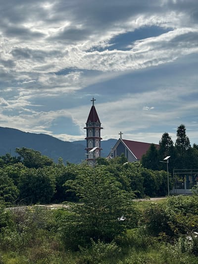 A picturesque view of a church is captured in the image, nestled amidst lush greenery and rolling hills. The church features a distinctive tiered bell tower topped with a red roof and a cross, standing prominently against a backdrop of dramatic, cloud-filled skies. Adjacent to the bell tower is the main body of the church, also with a red roof and a cross adorning its gable. The surrounding landscape is dense with various shades of green trees and bushes, with a road visible in the foreground. In the distance, a range of mountains forms a majestic silhouette against the sky. The overall impression is one of serene beauty, with the church serving as a focal point within a vibrant natural setting.