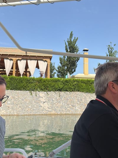 The image, when correctly oriented, depicts an outdoor scene under a clear blue sky, likely in Kuchtumgali, Uzbekistan. Two individuals are partially visible, engaged in a leisurely activity near a body of water.
In the right foreground, a man with grey hair is seen from behind and the side, wearing a black shirt with a red and white stripe on the collar. He appears to be looking towards the left of the frame. At the bottom left, the top of a second person's head is visible, wearing glasses and a striped top, also looking towards the scene.
To the right of the figures is a tranquil body of water, possibly a pond or canal, exhibiting reflections on its surface. Adjacent to the water is a path or border paved with small, light-colored pebbles or cobblestones.
Further into the mid-ground and background, a well-maintained green hedge separates the water feature from various architectural elements and foliage. On the left, a building with ornate, carved wooden columns is visible, featuring light-colored draped fabric, possibly awnings or shades, suggesting a sheltered area. This building displays intricate patterned tile work. In the distance, behind the hedges and trees, a tall, light-colored brick structure, resembling a minaret or tower, stands prominently against the clear blue sky. Scattered green trees add to the natural landscape.
The bright sunlight and clear sky indicate a pleasant day. The architectural style, with its patterned masonry and the tall tower, aligns with the provided location context of Uzbekistan, suggesting a historical or tourist site.