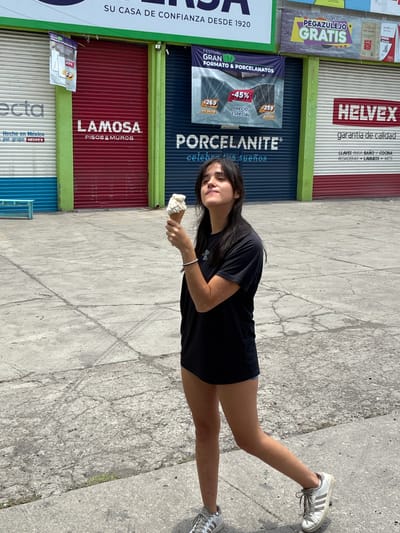 A young woman stands on a concrete sidewalk, holding an ice cream cone, in front of a row of storefronts with closed metal shutters. The storefronts are adorned with advertisements for brands such as Lamosa, Helvex, and Porcelanite. These appear to be stores that sell flooring, tile, plumbing supplies, and other home improvement goods. An older sign visible states: "Su Casa de Confianza Desde 1920." The text along with these brands places the location in Mexico. The advertisement for "Hecho en Mexico por grupo Helvex" suggests the city is in Tepeji de Ocampo, Mexico. The woman wears a black t-shirt and white sneakers. The sidewalk is cracked in places. The light suggests it is midday.