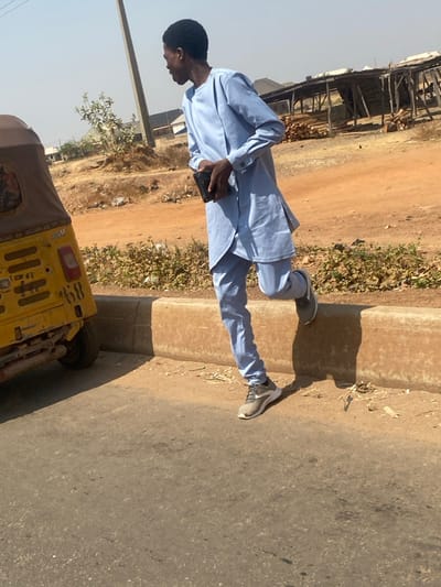 This image depicts an outdoor scene in Lapai, Nigeria, under bright, clear daylight. A lean, dark-skinned male, appearing to be a young adult, is positioned with his left foot on an asphalt road and his right foot elevated and resting on a low, light brown concrete curb. He is dressed in a light blue traditional tunic with long sleeves and matching trousers, complemented by light grey athletic shoes. In his left hand, he holds a dark, rectangular object. His head is turned to the left, and his gaze is directed off-frame.
To the immediate left of the male, the rear-left section of a yellow auto-rickshaw is partially visible on the asphalt, displaying the number "68" on its back panel. Beyond the concrete curb, a dirt shoulder with sparse, dry vegetation extends into the mid-ground. The background features a tall, slender utility pole, several low-rise buildings with light-colored walls and dark roofs, and a wooden skeletal structure with stacked timber to the far right. The visible ground in the background consists primarily of dry, reddish-brown earth. Distinct shadows are cast on the ground, indicative of a high sun angle.