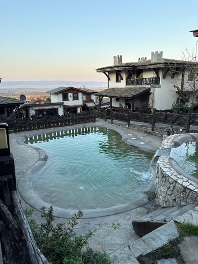 The image depicts an outdoor pool and surrounding structures in Medovo, Bulgaria. A large, irregularly shaped pool with light-colored water dominates the center. A smaller, elevated pool with a waterfall feature feeds into the larger pool. The pools are surrounded by stone and concrete.
Adjacent to the pool are buildings constructed in a rustic style, featuring white walls, dark wood accents, and stone roofing. A wooden fence borders the pool area. In the background, a valley landscape stretches towards a horizon with a gradient sky. The image is shot from an elevated perspective, possibly from a balcony or deck.