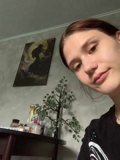A young woman is taking a selfie in an indoor setting, possibly her home. She has medium-brown hair pulled back, and her face is turned towards the camera. She is wearing a black t-shirt with a graphic design. In the background, a painting of a silhouette against a celestial backdrop hangs on a textured gray wall. To the right of the painting is a decorative artificial tree with green and white leaves. On a dark wooden surface in the foreground, there are various items including a glass jar, some boxes, and a small mug with flowers. The lighting appears to be ambient indoor light.
