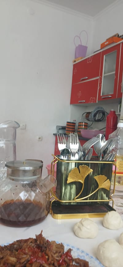 The image depicts a kitchen scene, likely in Mihaylovka, Kyrgyzstan. In the foreground, a plate of food is visible, with a glass teapot to its left. A utensil holder, decorated with gold leaf motifs, is centrally placed, containing forks and spoons. Further back, a red cabinet with glass doors is mounted on the wall. The environment suggests a domestic setting, possibly a breakfast or meal preparation area. There are no people visible in the frame. The lighting suggests it is daytime.