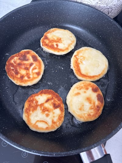 The image shows six circular, dough-based food items frying in a black non-stick pan with white speckles. The items display various levels of golden-brown caramelization on their surfaces, indicating they are in the process of being cooked. A thin layer of cooking oil is visible in the pan. A portion of a black induction stovetop control panel, featuring white etched symbols including a timer icon and plus/minus buttons, is visible in the lower left. In the upper right background, a segment of a white ceramic or porcelain object with black floral or leafy patterns is partially visible. No individuals or direct interactions are present. The indoor environment lacks any specific visual cues that would enable the identification of its geographic location, including the city of Zmeinyi, Russia.