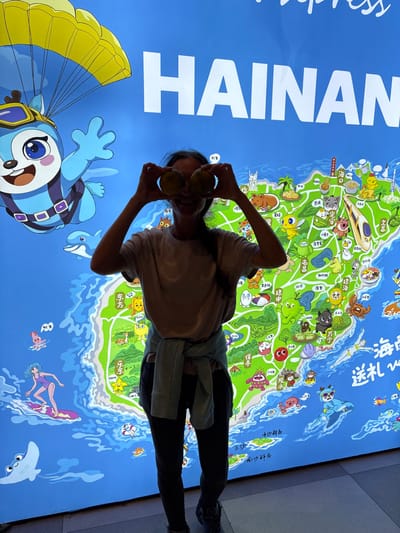The image displays a single adult female standing indoors in Tianya District, China, positioned directly in front of a large, brightly illuminated wall display. The display features a stylized, vibrant green and blue cartoon map of Hainan Island, with the word "HAINAN" prominently spelled in large white capital letters across the top. The map is adorned with numerous cartoon illustrations, including a blue, deer-like character parachuting in the upper left, a dolphin, and a female character surfing. The female subject, with visible dark hair, wears a light-colored short-sleeved t-shirt, dark fitted pants, and dark athletic shoes; a light-colored long-sleeved garment is tied around her waist. She is holding two round, yellowish-brown objects, possibly fruits, directly in front of her eyes, obscuring them. The subject is smiling with her mouth open. The floor beneath her consists of light-toned tiles.