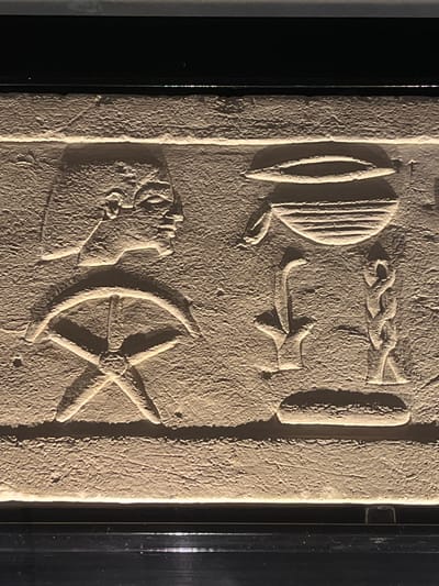The image shows a close-up of a bas-relief carving in an ancient Egyptian style. On the left, a profile of a human head, possibly male, is depicted wearing a headdress. Below the head is a bow and arrow symbol, with the bowstring forming a star-like shape. To the right of the human head and bow, there is a collection of hieroglyphic symbols. These include a large oval shape with horizontal lines inside, suggestive of a basket or tray, and below it, two smaller symbols, one resembling a stylized plant or reed and the other a coiled rope or knot. The carving is illuminated from the bottom, creating shadows that highlight the texture of the stone and the depth of the relief. The background is dark, suggesting the carving is set within a display or architectural feature. The carving is located in Cologny, Switzerland.