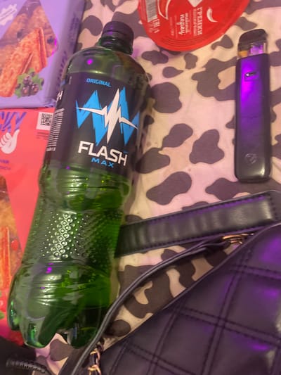 The image depicts a still life arrangement on a patterned surface, possibly a bedspread with a cow-print or similar design. The focal point is a large, green plastic bottle of "FLASH MAX" energy drink, positioned prominently in the foreground, tilted to the right. To the right of the bottle, a dark purple vaping device with a glowing light is visible. In the background, partially obscured, are packages of food items, including what appears to be crackers and a red, circular food container. The lighting suggests a dim environment, with a purple hue cast over parts of the scene, possibly from an artificial light source. There are no people or discernible actions depicted in the image. The location in the city of Zmeinyi, Russia cannot be determined from the visual information provided.