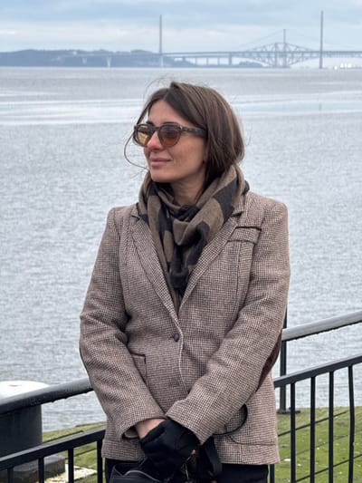 An adult female stands outdoors in the city of Blackness, Scotland, United Kingdom, facing right with a slight smile. She wears dark-framed sunglasses with amber-tinted lenses, a brown and dark grey striped scarf, a light brown and beige houndstooth blazer featuring dark elbow patches and piping, and black gloves. Her hands are clasped in front of her, holding a black object or bag. The background features the calm grey water of the Firth of Forth under an overcast sky, with three distinct Forth Bridges spanning the horizon from left to right: a cable-stayed bridge, a suspension bridge, and a cantilever railway bridge. In the foreground, a black metal railing runs along the bottom, and a dark bollard is partially visible on the left, resting on a moss-covered stone or concrete edge.