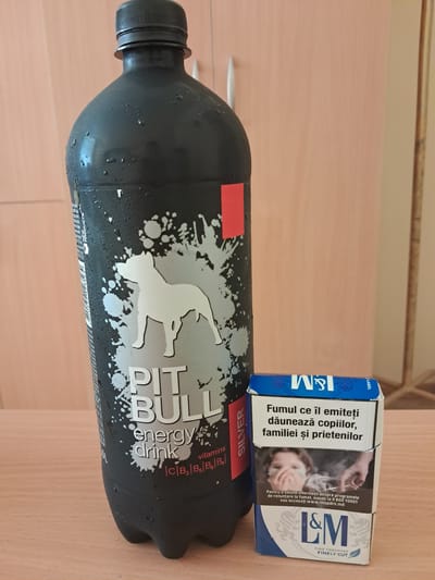 The image shows a close-up, overhead view of a dark gray "Pit Bull Energy Drink" bottle lying on its side next to a pack of L&M cigarettes. The bottle has a distressed, splatter-like design with a silver silhouette of a bull. The label clearly reads "PIT BULL energy drink" and "SILVER." There are water droplets on the bottle, suggesting it is cold. The cigarette pack is blue and white, with the "L&M" logo prominently displayed. The pack also contains Romanian text warning about the dangers of smoking, stating "Fumul ce îl emiteți dăunează copiilor, familiei și prietenilor" (The smoke you emit harms children, family and friends). The background is a light-colored, textured surface, likely a table. The lighting suggests it is daytime. The overall scene implies a casual, possibly personal setting.