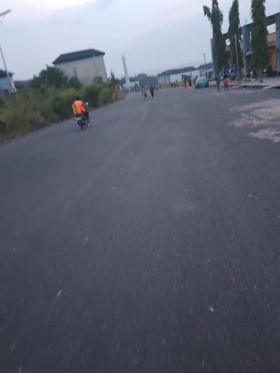 The image depicts a paved road in Ogboko, Nigeria, likely a street within a residential or commercial area. The road is gray and shows signs of wear.
A person on a motorcycle with an orange backpack is driving on the left side of the road, moving away from the camera. Several individuals are walking on the right side of the road, closer to the buildings.
There are buildings on either side of the street. On the left, there is a large, light-colored building behind some vegetation. In the background, a minaret is visible. On the right, there is a building with tall, narrow pillars or columns. A car is parked in front of this building.
The sky is overcast and the light appears soft, suggesting either early morning or late afternoon. The image shows the intersection of vehicular and pedestrian traffic.