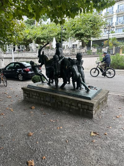 The image captures a detailed outdoor scene in Vevey, Switzerland, specifically on the Quai Ernest-Ansermet, facing the Hotel des Trois Couronnes.

In the foreground, a large, dark bronze sculpture is prominently displayed on a multi-tiered stone base. The sculpture, identified as "La Joie de Vivre" by Jean-Pierre Zaugg, depicts approximately six to seven nude or partially draped youthful human figures dynamically interacting with a horned quadruped, resembling a ram or ibex. One figure appears to pull the animal's head, another is seated on its back, and others push or engage with its body. The ground surrounding the sculpture is a light grey gravel path, scattered with numerous brown fallen leaves. A portion of a bicycle, visible on the far left, includes a red frame and front wheel.

Behind the sculpture, a paved road stretches horizontally. A black two-door coupe is stationary on the left side of the road, oriented away from the viewer. On the right side of the road, a male individual wearing a dark jacket, light trousers, and a dark helmet rides a black e-bike from right to left, with his head turned towards the left.

In the midground, a stone wall with black metal railings separates the road from an elevated terrace featuring white outdoor umbrellas and tables, indicative of a cafe or restaurant. Lush green trees with broad leaves are abundant throughout the midground and background. The background is dominated by a grand, light-colored multi-story building with numerous balconies and windows, consistent with the architecture of the Hotel des Trois Couronnes. A decorative black streetlamp with multiple luminaires stands in front of this building. The scene is illuminated by diffused natural daylight.