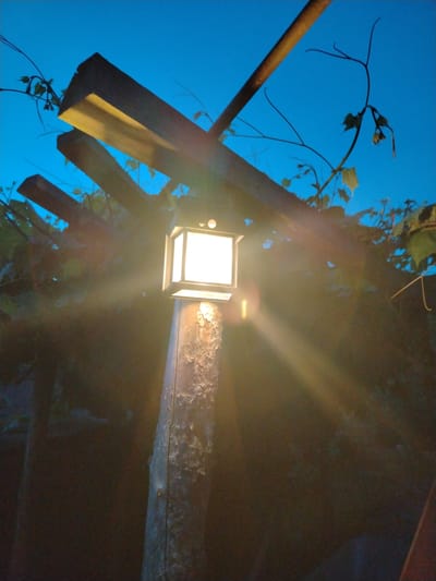The image shows a solar-powered lantern attached to a wooden post, illuminated against a dark blue twilight sky. The lantern emits a bright light that creates visible rays and lens flare. The post is rough and weathered, with peeling paint. Above the post, a wooden pergola with parallel beams extends across the frame. Green vines with leaves are growing around and on the pergola, suggesting an outdoor setting, possibly a garden or backyard. The scene is set in the village of Asnezhytski Sel'ski Savet, Belarus.