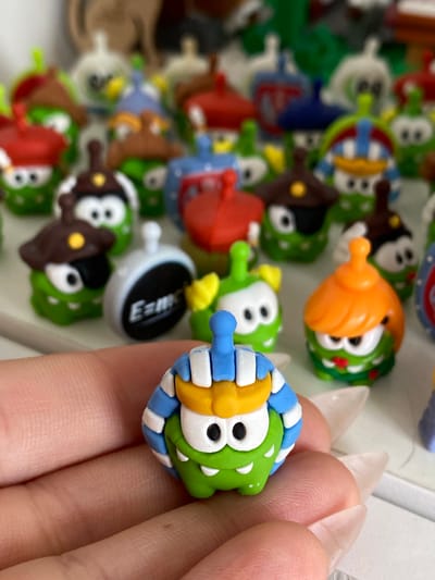 A close-up, shallow depth-of-field shot shows a person's hand holding a collection of small, green, cartoonish figures. The figures have large white eyes with black pupils and wide mouths. Many are wearing different colored hats and accessories. The figure in the foreground is green with white, blue, and yellow striped headwear. In the background, a blurred array of similar figures is visible, some with red, blue, or brown hats. One figure in the middle ground has a gray, circular accessory with "E=mc²" written on it. The overall aesthetic is playful and colorful, suggesting a collection of toys or trinkets. No discernible urban or natural environmental features are present, making location identification within Ostrovki, Russia impossible from the visual information provided.