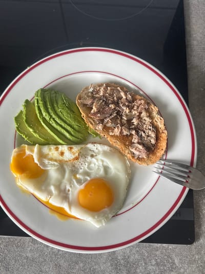 This image, captured in Tielrode, Belgium, displays a prepared meal presented on a white circular plate featuring a red outer rim. The plate contains two sunny-side-up fried eggs with bright yellow yolks, multiple fanned-out slices of green avocado, and a single piece of toasted bread topped with flaked, light-brown protein, consistent with fish. A silver-colored, four-tined fork rests on the right side of the plate, partially overlapping it. The plate is positioned on a dark, reflective surface, consistent with an induction stovetop, with a lighter grey, speckled countertop visible to the right. No individuals are present in the scene, and no active interactions are depicted.
