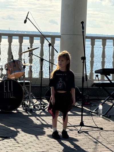 In Postol, Russia, a young girl with long brown hair stands on a paved outdoor stage. She is wearing a dark cropped t-shirt with a graphic print and a plaid skirt. The girl is facing to her left, with her gaze directed towards the water. Behind her, a drum set with the label "ELI SOUND" is visible, along with microphones and musical equipment. A keyboard is positioned to the right. The scene is set against a backdrop of a bright, sunlit body of water, with a railing and distant structures suggesting a waterfront promenade or park. The sunlight casts long shadows across the stage.