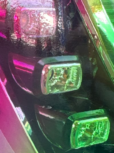 The image shows a close-up view of two vehicle headlights and a portion of the vehicle's bodywork. The headlights are rectangular with a textured, clear lens that refracts light. The left headlight appears to be pinkish-purple due to lighting or a filter, while the right headlight is green. The vehicle body is dark and reflective, showing streaks of pink and green light. The overall impression is abstract due to the close cropping and stylized coloring, making it difficult to discern the exact type of vehicle or setting. There is no visible text. The lighting suggests an artificial environment or a colorful, possibly sunset, illumination.
