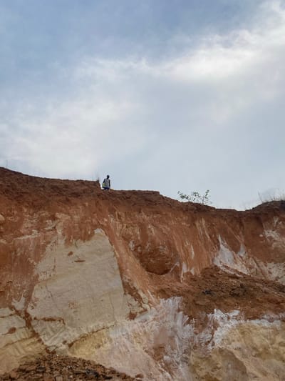 An outdoor scene in Asaga Amangwu, Nigeria, displays a prominent, multi-layered earthen cliff under a predominantly overcast sky. A single individual stands on the uppermost edge of the cliff, positioned left of the image's vertical center, facing towards the right. The person is dressed in a light-colored upper garment and dark lower garment, with a light-colored object visible at their left side. The cliff face exhibits clear geological stratification: a thick upper layer of reddish-brown soil transitions into a substantial lower section of exposed pale yellow to off-white earth, likely composed of clay or sand. Visible erosion patterns, including vertical white streaking, mark the reddish earth. Sparse, dry vegetation is present along the top edge, with a small leafy plant situated further right on the crest. An accumulation of reddish-brown soil and small rocks forms a debris pile at the base of the cliff on the lower left. The sky is largely covered by diffuse grey and white clouds.