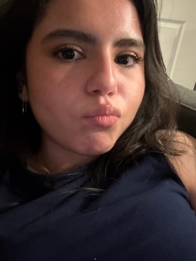 A close-up selfie of a woman with dark hair and brown eyes. She has full lips pursed in a kiss expression. Her eyebrows are thick and well-defined, and her eyelashes are long and dark. She is wearing a dark blue shirt. The background shows a blurry white door. The specific location in College Station, United States of America cannot be determined from this image.