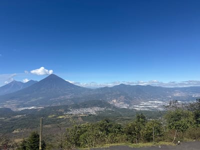 A wide shot reveals a landscape of mountains under a clear blue sky. Two prominent volcanic peaks dominate the scene, one with a wisp of cloud beside it. Patches of green forest and clustered settlements are visible in the lower valleys. Other mountains stretch along the horizon, fading slightly with distance. In the foreground, trees and foliage line what appears to be a roadside vantage point.