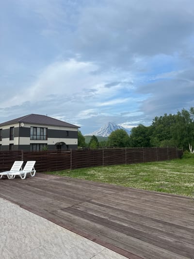 The image depicts a modern two-story house with a dark brown, pitched roof, featuring horizontal bands of dark grey and off-white on its exterior. The house has numerous windows, some tinted, and a satellite dish mounted on the upper facade. In front of the house, a dark brown wooden fence encloses a grassy yard dotted with small white flowers. Two white lounge chairs are positioned on a dark wooden deck in the foreground. Beyond the fence and the house, a majestic, snow-capped mountain, possibly a volcano, dominates the background under a cloudy sky. Lush green trees surround the mountain and extend to the right side of the frame. The scene appears to be set in a semi-rural or suburban area, likely in Yuzhnyye Koryaki, Russia, given the prominent volcanic landscape. There are no people or animals visible in the image.