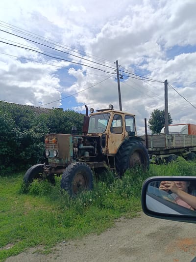 An abandoned tractor, painted beige and teal, sits in tall green grass beside a wooden trailer filled with cargo covered by an orange tarp. The tractor is weathered and rusty, with large, treaded tires. Behind the tractor, a dense line of green foliage borders a building with a grey, corrugated roof. Overhead, power lines stretch across a partly cloudy sky with fluffy white clouds. In the foreground, the reflection in a car's side mirror shows a person wearing a pink shirt and holding a phone. The scene is set on a dirt road in the city of Kamennoye, Russia.