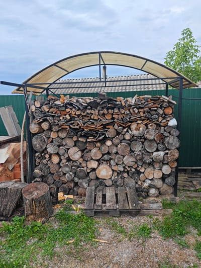 The image depicts a woodpile stored under a metal-framed awning with a translucent fabric roof. The woodpile is substantial, composed of various sizes of logs and split wood, neatly stacked against a green corrugated metal fence. The logs are cut to different lengths and diameters, with visible grain patterns and bark textures. Some of the wood appears to be seasoned. In front of the woodpile, there are two large tree stumps, one with prominent bark texture and the other with a smooth, cut surface. Partially visible to the left of the woodpile are some construction materials, including bricks and flat concrete-like slabs. The ground in front of the woodpile is covered with gravel and patches of green grass and weeds. The sky above is overcast with a cloudy, pale blue hue. The overall scene suggests an outdoor storage area, possibly in a rural or semi-rural setting. There are no people visible in the image. The city of Песчанка, Russia, is not discernible from the visual information provided in the image.