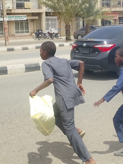 The image depicts a street scene in Gyadi Gyadi Arewa, Nigeria, under bright daylight with strong shadows. Two young males are in motion on an asphalt road. The primary figure, a young male wearing a charcoal grey short-sleeved tunic and matching trousers with light-colored sandals, runs from left to right, carrying a full light-yellow plastic bag in his left hand. His head is turned to the right. To his right, partially visible, a second young male in a blue top and trousers extends his left arm forward with an open hand, reaching towards the first boy. In the background, a black Toyota Corolla sedan is visible on the road. Across a curb with black and white stripes, a light-colored commercial building with a visible sign for 'Qisol Ventures Ltd' stands. Several motorcycles, some fitted with delivery boxes, are parked in front of this building. Two large palm trees are situated between the building and the road.