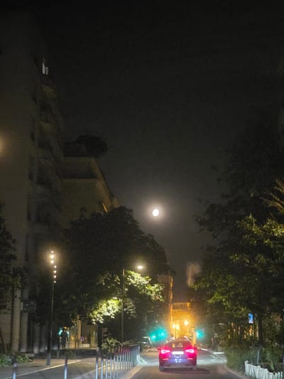 An eye-level, medium shot captures a street scene at night in Montrouge, France. The full moon is visible in the dark, hazy sky, casting a soft glow. A red car is the primary subject in the foreground, its taillights illuminated as it drives down the road. The street is lined with trees that cast dark shadows, and lampposts provide artificial light. Buildings flank the street, their windows glowing with interior light. The road surface is asphalt, with white lane markings. A sidewalk is visible on the left, with a row of bollards separating it from the street. The overall atmosphere is calm and slightly mysterious, with the limited light sources creating pockets of illumination and deep shadows.