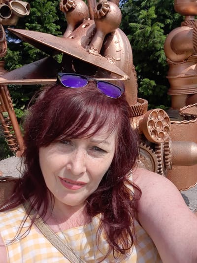 In a close-up, selfie-style shot, a middle-aged woman with auburn hair and freckles smiles at the camera. She wears purple sunglasses perched on her head and a yellow and white checkered dress. A silver chain necklace is visible. She is positioned in front of a large, metallic sculpture with copper-toned components, including gears and pipes. In the background, lush green pine trees are visible. The scene appears to be outdoors, possibly in a park or garden setting, given the presence of foliage and what looks like a stone or concrete surface in the foreground. The woman's arm is also visible, resting on what seems to be a stone surface. The overall impression is a candid outdoor portrait with a distinctive artistic backdrop.