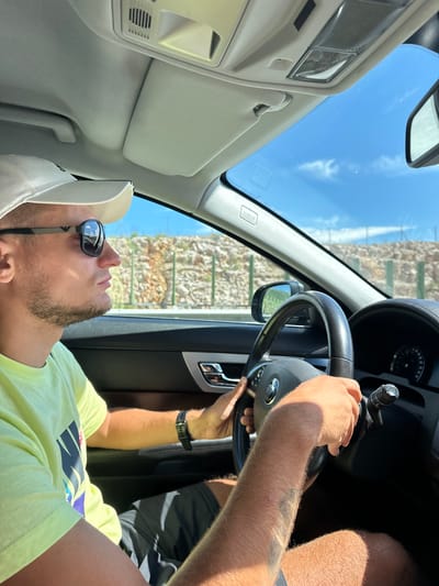 A man wearing a white baseball cap, sunglasses, and a neon yellow t-shirt is driving a Jaguar in Pelev Brijeg, Montenegro. The car's interior is visible, including the steering wheel, dashboard, and part of the ceiling. The man's hands are on the steering wheel. The exterior scenery shows a rocky hillside with sparse vegetation and a clear blue sky. The sun is shining brightly, casting shadows inside the car.