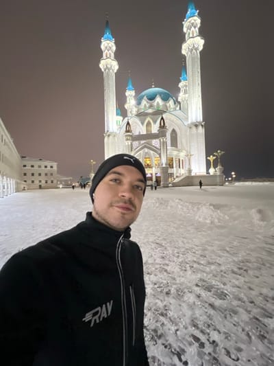 A light-skinned adult male, approximately mid-20s to mid-30s, is depicted in a selfie taken at night. He wears a black beanie with a white Puma logo on the front, a black zip-up athletic jacket with a white "RAV" logo on the left chest, and has short dark hair with a light beard or stubble. He looks directly at the camera with a neutral expression.
The background features the prominently illuminated Kul Sharif Mosque, located within the Kazan Kremlin in Kazan, Republic of Tatarstan, Russia. The mosque is characterized by its white facade, four tall, slender minarets topped with pointed blue finials, and multiple blue, ribbed domes, with a large central dome. The entire structure is brightly lit with white and blue artificial lights, accentuating its architectural details against the dark night sky.
To the left of the mosque, a multi-story building with a white facade and numerous windows is visible, partially illuminated. Further left, a white colonnaded structure can be seen. The ground throughout the scene is entirely covered in a thick, uneven layer of white snow, showing some footprints and disturbed areas. The sky is dark, indicating nighttime, with a subtle reddish-orange luminescence near the horizon, possibly from city light pollution. Several small, indistinct figures are visible walking on the snow-covered grounds in the middle distance near the mosque and adjacent buildings.