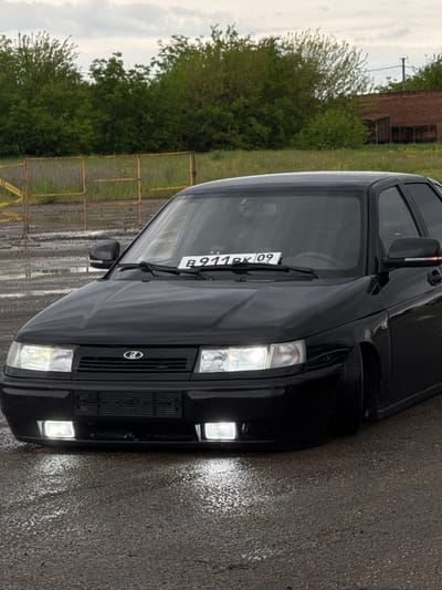The image shows the front of a black car, specifically a Lada or similar model, with its license plate visible. The license plate reads "B911PK09". The car is parked on wet asphalt. In the background, there's a blurred view of green foliage, possibly trees and shrubs, along with what appears to be a rusty metal structure or building. The sky is overcast. Considering the "09" on the license plate, the car may be from the Republic of Kalmykia, Russia. While a specific location cannot be definitively confirmed to be Pavlovskaya, Russia from the image alone, the scene suggests a rainy day in a slightly industrial or semi-rural area within Russia, given the car model, license plate, and background elements. The car lights are on. There is an orange structure in the background.