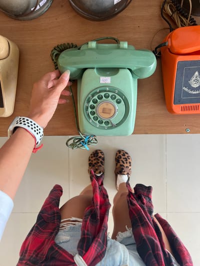 In Tambon Phraek Ha, Thailand, a person wearing leopard-print slippers and a red and black plaid shirt tied around their waist over distressed denim shorts is standing on a tiled floor, facing a wooden surface. The person's left arm, adorned with a white Apple Watch band, is reaching towards a vintage green rotary phone. Another vintage orange rotary phone is situated to the right of the green phone. The phones are on a wooden shelf, with other vintage items visible in the background. The scene is a top-down perspective.