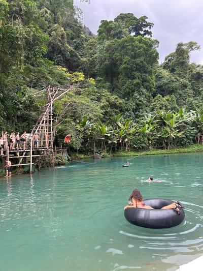 A scenic view features a lush green environment and light turquoise water. A wooden structure with several people visible on it sits near the water's edge, with one person swinging from a rope. In the water, several individuals are swimming, including a woman relaxing in an inner tube. The water appears calm and inviting against the backdrop of dense trees and foliage. The overcast sky complements the serene atmosphere.