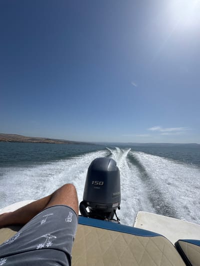 The image shows the rear of a boat traveling across the Sea of Galilee, a large freshwater lake in northern Israel. The coordinates 32.849, 35.538 place the location near Ginosar, a village on the western shore of the lake. The scene is captured from the perspective of a person seated in the back of the boat. The boat is moving forward, creating a wake behind it. The person is wearing grey shorts with a white pattern, and the boat has a tan, quilted, cushioned seat. A Yamaha outboard motor with a "150" designation is visible behind the person. The sun is shining brightly, creating glare on the water, and a clear blue sky is visible above. The shoreline can be seen in the distance.