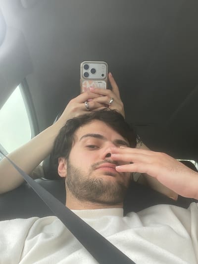 A male person is inside a vehicle, likely a car, and is seen in a selfie pose. The car's interior, including the roof and window, is visible. He's wearing a white sweater. The man is wearing a seatbelt. He is holding a phone in his left hand. The phone has a clear case with a design on it. The background indicates a cloudy or overcast day. The location cannot be determined.