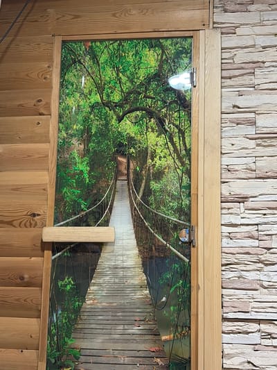 A full-shot, eye-level photograph shows a wooden door with a vibrant mural of a suspension bridge in a lush, green forest. The door is framed by wooden paneling on the left and stacked stone on the right. A small, silver door hinge is visible on the right side of the door frame. The mural depicts a wooden plank bridge with rope railings, winding into a dense forest with sunlight filtering through the trees. Fallen leaves are scattered on the bridge. The location is not specified as being in the city of Субханкулово, Russia.