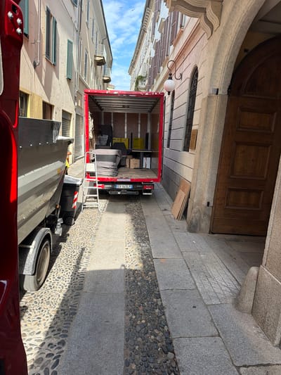 This is a photograph taken outdoors in Novara, Italy, during the day. The scene appears to be a narrow street with buildings on either side, possibly an urban setting. The main focus of the image is a red moving truck backed up against a building with a large wooden door. The truck is partially open, revealing furniture and boxes inside, suggesting a move or delivery is in progress. A ladder is extended from the truck to the ground.
To the right of the truck, the ground is paved with granite slabs and small pebbles. In the foreground, the edge of a red vehicle is visible, and to the far right, a spare tire is resting on the ground. The buildings flanking the street have architectural details, including balconies and shuttered windows, typical of Italian architecture. The sky above is blue with some clouds and birds flying.
There is some visible text on the truck: "Iveco DAILY" and a license plate "ER 943LP". There is also text on some of the boxes inside the truck, but it is not legible. The lighting suggests it is daytime, likely sunny, casting shadows on the ground. The overall mood is one of activity and transition, given the presence of the moving truck and its contents.